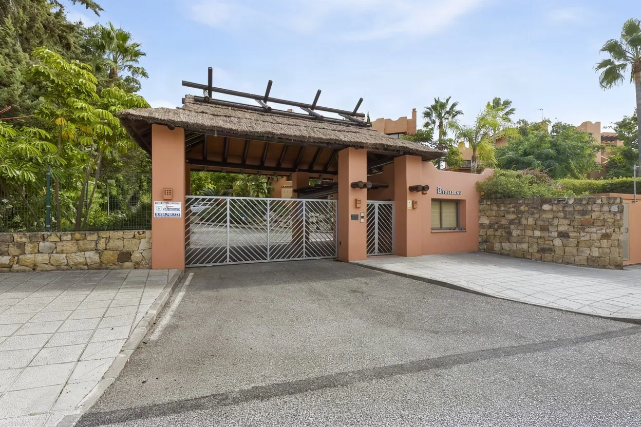 Entrance gate of a residential complex with terracotta walls, a thatched canopy and palm trees, edited with Stager AI