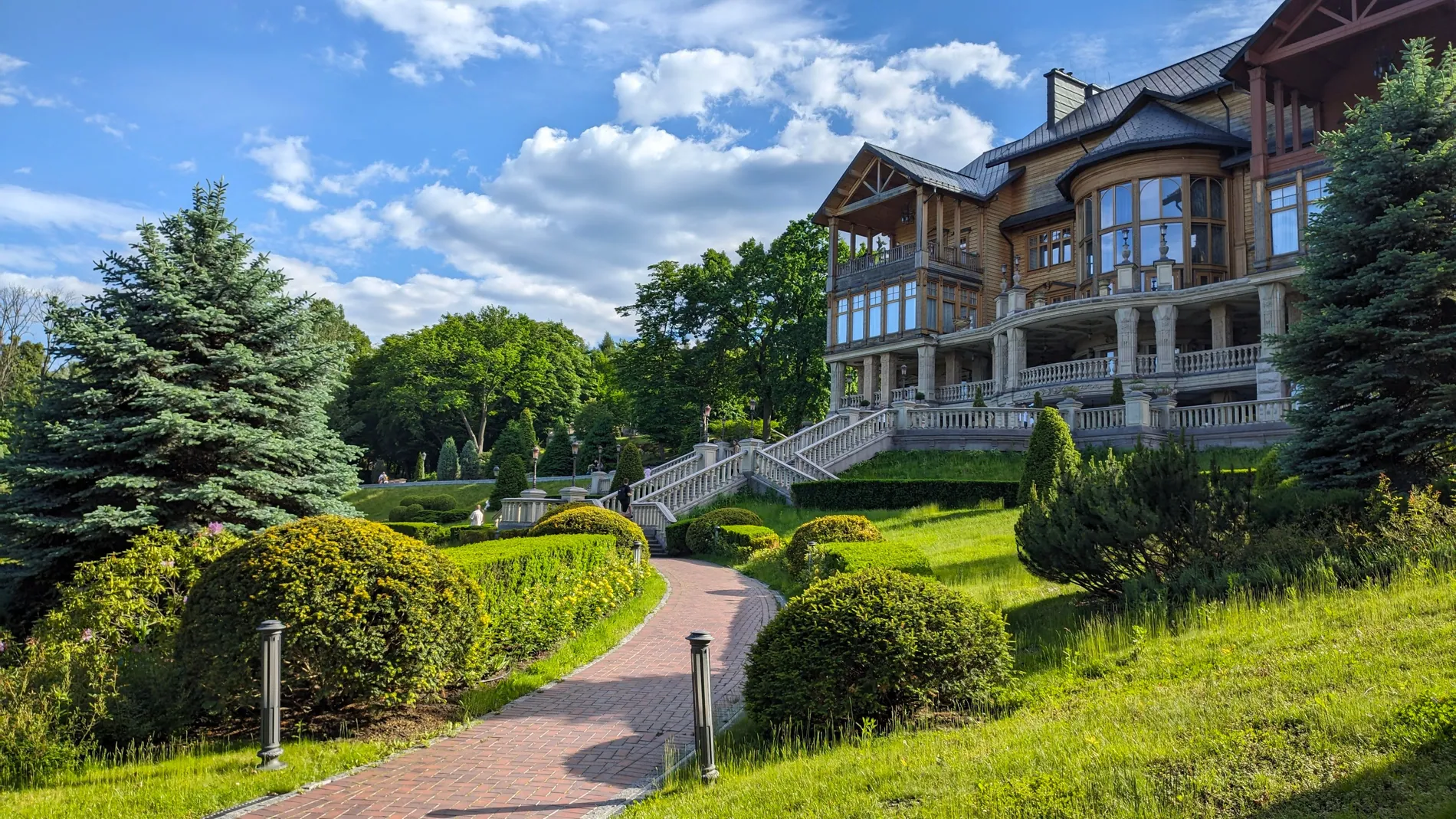 Large house on a hillside garden under a stormy sky, replaced by a bright blue summer sky