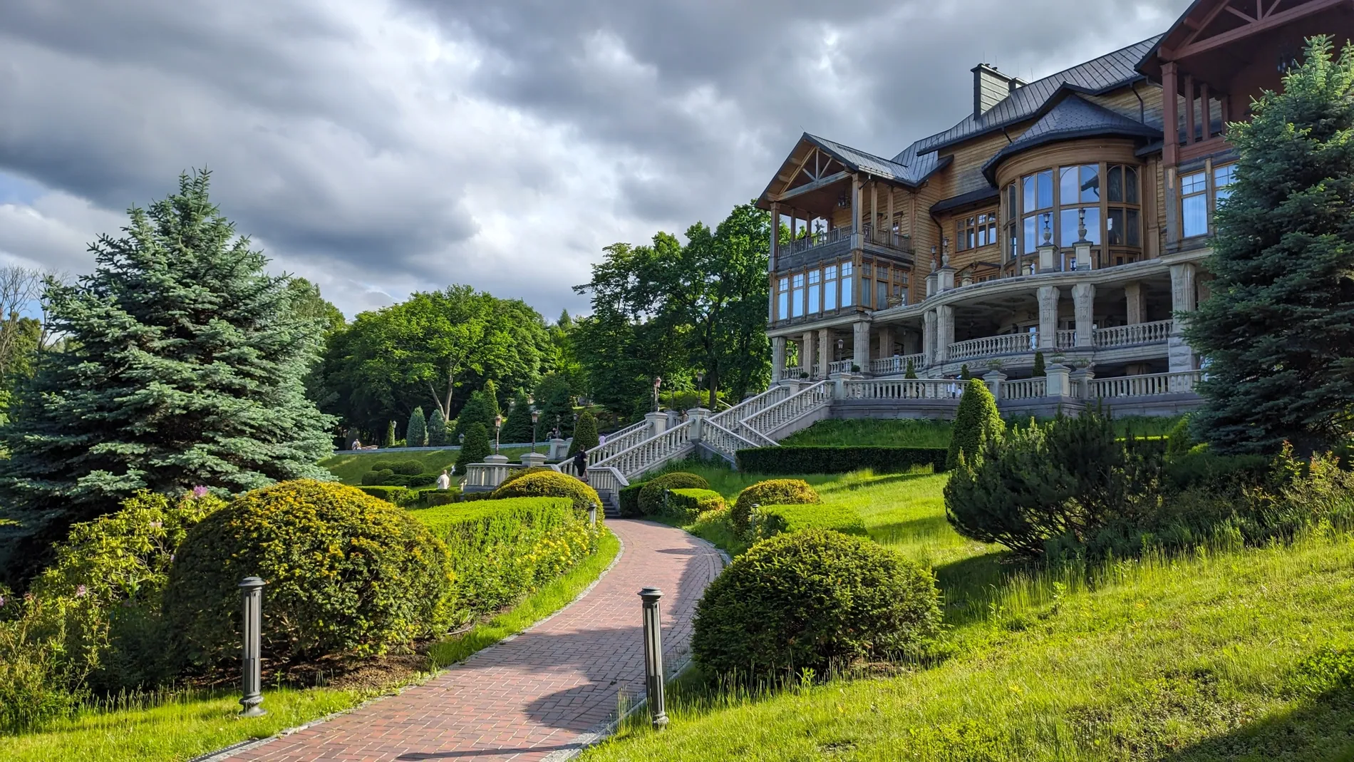 Large house on a hillside garden under a stormy sky, replaced by a bright blue summer sky