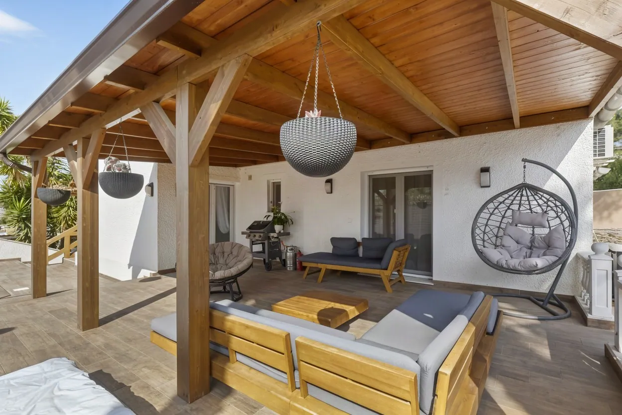 Covered wooden terrace of a white Mediterranean house with outdoor sofas, a hanging egg chair and pendant planters